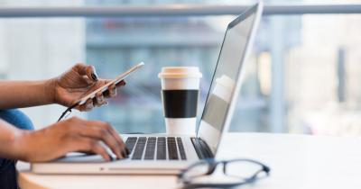 Person sitting at a laptop typing with right hand and holding mobile phone in left hand
